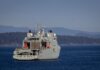 A military warship navigates through open waters with a coastline backdrop on a clear day.