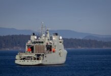 A military warship navigates through open waters with a coastline backdrop on a clear day.