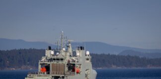 A military warship navigates through open waters with a coastline backdrop on a clear day.