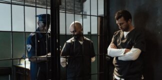 A police officer stands by two prisoners in jail uniforms behind bars, indoors.