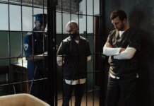 A police officer stands by two prisoners in jail uniforms behind bars, indoors.