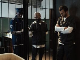 A police officer stands by two prisoners in jail uniforms behind bars, indoors.