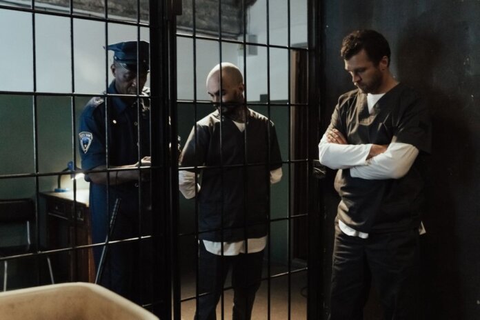 A police officer stands by two prisoners in jail uniforms behind bars, indoors. A police officer stands by two prisoners in jail uniforms behind bars, indoors.