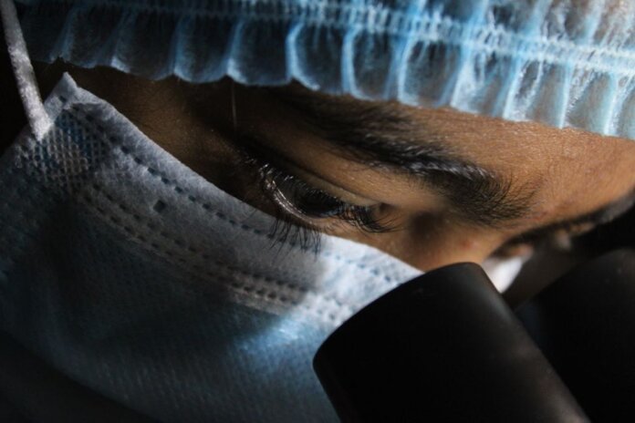 A scientist in protective gear intensely looking through a microscope.