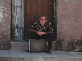 A senior man in a patterned shirt sits with a suitcase on a rustic doorstep.