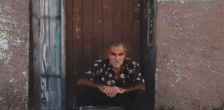 A senior man in a patterned shirt sits with a suitcase on a rustic doorstep.