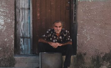 A senior man in a patterned shirt sits with a suitcase on a rustic doorstep.