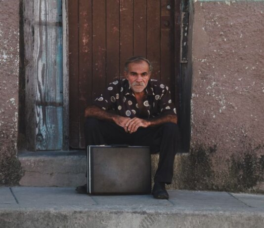 A senior man in a patterned shirt sits with a suitcase on a rustic doorstep.