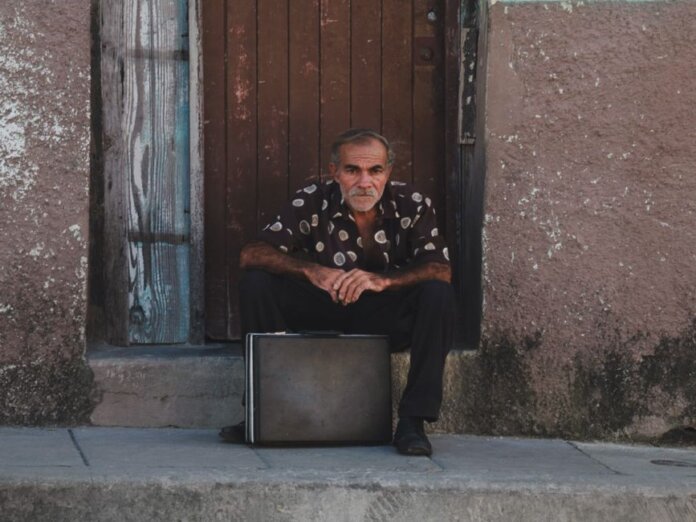A senior man in a patterned shirt sits with a suitcase on a rustic doorstep. A senior man in a patterned shirt sits with a suitcase on a rustic doorstep.