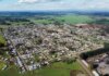 Aerial photo of a rural town surrounded by green fields under a cloudy sky, showcasing its layout.