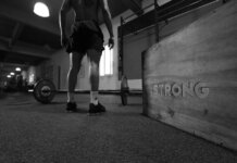 Black and white photo of a man in a gym with barbell and 'Strong' box.