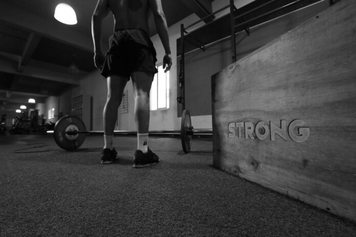 Black and white photo of a man in a gym with barbell and 'Strong' box. Black and white photo of a man in a gym with barbell and 'Strong' box.