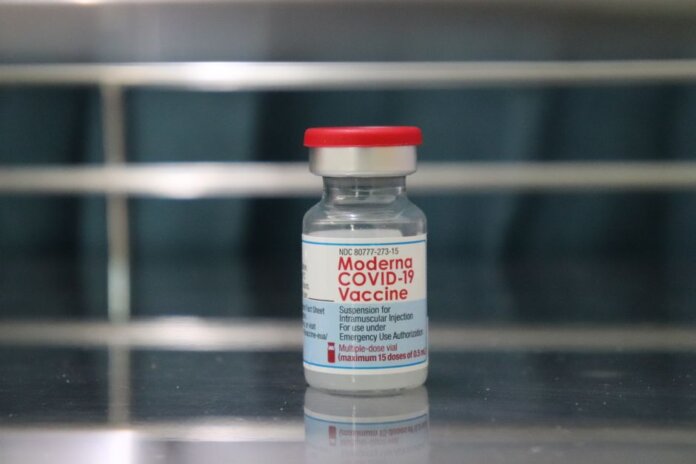 Close-up of a Moderna COVID-19 vaccine vial on a reflective metal surface.