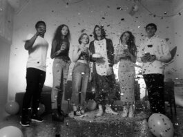 Group of friends celebrating indoors with confetti and balloons in a black and white setting.
