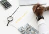 Hands folded over stock market documents with calculator and cash on desk.