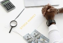 Hands folded over stock market documents with calculator and cash on desk.