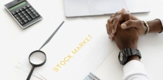 Hands folded over stock market documents with calculator and cash on desk.