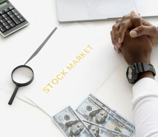 Hands folded over stock market documents with calculator and cash on desk.