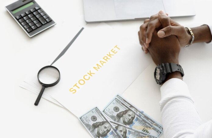 Hands folded over stock market documents with calculator and cash on desk. Hands folded over stock market documents with calculator and cash on desk.