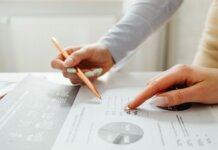 Hands holding a pencil, analyzing charts and data sheets, suggesting teamwork in an office setting.