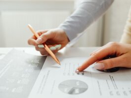 Hands holding a pencil, analyzing charts and data sheets, suggesting teamwork in an office setting.