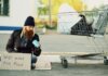 Homeless man sitting beside shopping cart with a sign "Will work for food."