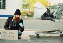 Homeless man sitting beside shopping cart with a sign "Will work for food."