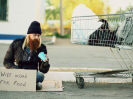 Homeless man sitting beside shopping cart with a sign "Will work for food."