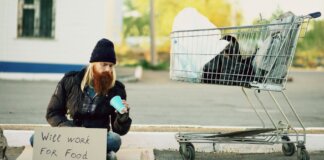 Homeless man sitting beside shopping cart with a sign "Will work for food."