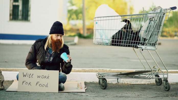 Homeless man sitting beside shopping cart with a sign "Will work for food."