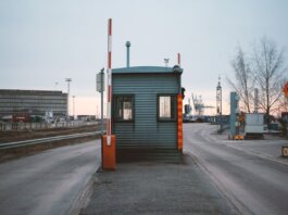 Industrial checkpoint and barrier at port under cloudy sky with cranes in the background.