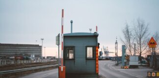 Industrial checkpoint and barrier at port under cloudy sky with cranes in the background.