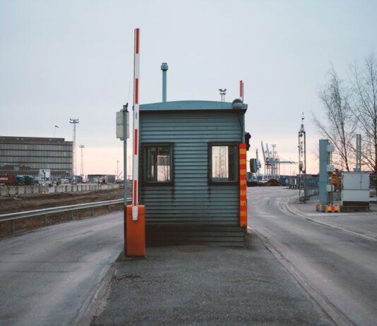 Industrial checkpoint and barrier at port under cloudy sky with cranes in the background.