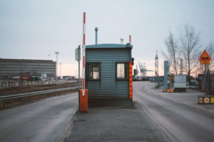 Industrial checkpoint and barrier at port under cloudy sky with cranes in the background. Industrial checkpoint and barrier at port under cloudy sky with cranes in the background.
