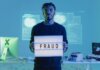 Man holding a 'FRAUD' sign in a tech setting, symbolizing cybersecurity threats.