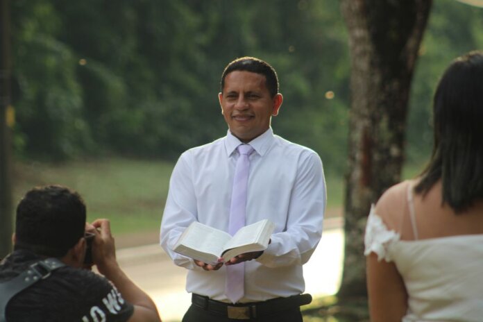 Pastor conducts a ceremony outdoors holding an open book with wedding participants.