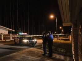 Police officers at a nighttime crime scene with a police car and caution tape under streetlights.