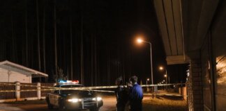 Police officers at a nighttime crime scene with a police car and caution tape under streetlights.