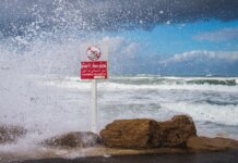 Powerful waves crash against the rocks with 'Swimming Prohibited' sign in Tel Aviv beach scene.