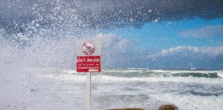 Powerful waves crash against the rocks with 'Swimming Prohibited' sign in Tel Aviv beach scene.