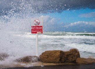 Powerful waves crash against the rocks with 'Swimming Prohibited' sign in Tel Aviv beach scene.