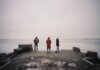 Three individuals are standing on a pier overlooking a frozen lake on a cloudy winter day.
