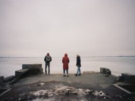 Three individuals are standing on a pier overlooking a frozen lake on a cloudy winter day.