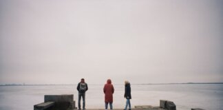 Three individuals are standing on a pier overlooking a frozen lake on a cloudy winter day.