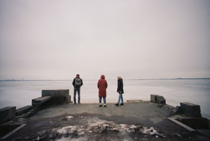 Three individuals are standing on a pier overlooking a frozen lake on a cloudy winter day.