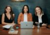 Three professional women sitting at a wooden desk in an office with laptops and notebooks.