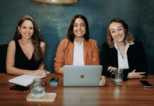 Three professional women sitting at a wooden desk in an office with laptops and notebooks.