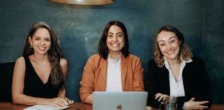 Канада выделит средства на поддержку женщин-предпринимателей Three professional women sitting at a wooden desk in an office with laptops and notebooks.