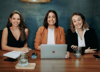 Three professional women sitting at a wooden desk in an office with laptops and notebooks.