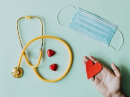 Top view of crop anonymous person hand with red paper heart on table with stethoscope and medical mask for coronavirus prevention